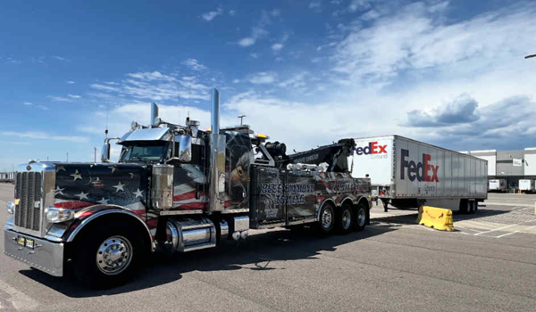 Tow truck with patriotic flag design towing a FedEx trailer on a paved lot under a blue sky.