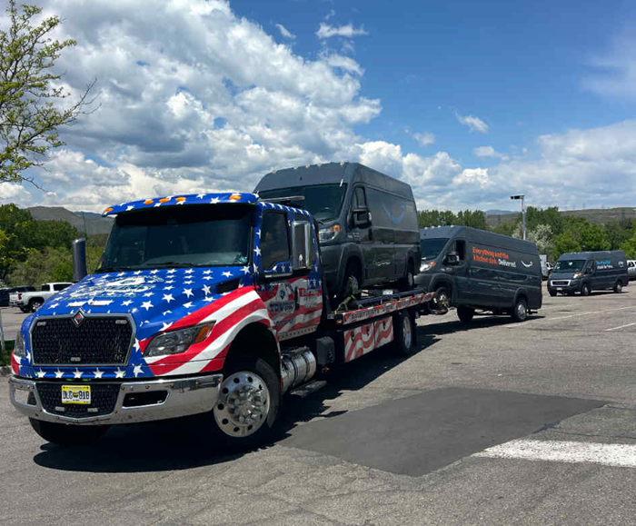 Tow truck with American flag design carries two vans in a parking lot on a sunny day.