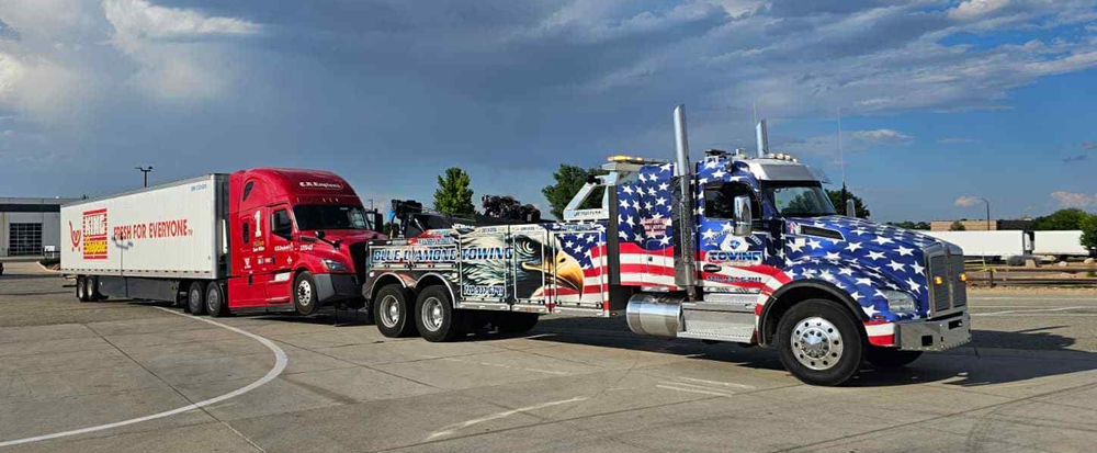 A tow truck with American flag design towing a red semi-truck on a gray paved area.