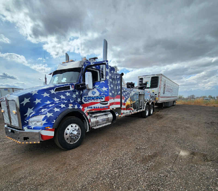 Tow truck with patriotic American flag design towing a trailer on a gravel lot under a cloudy sky.