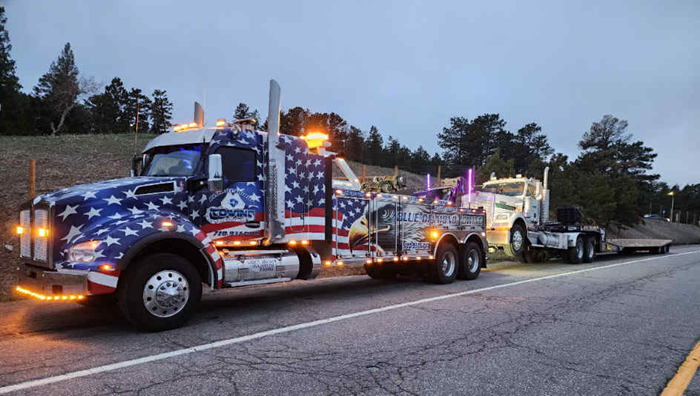 Tow truck with an American flag design towing another truck on a road at dusk.
