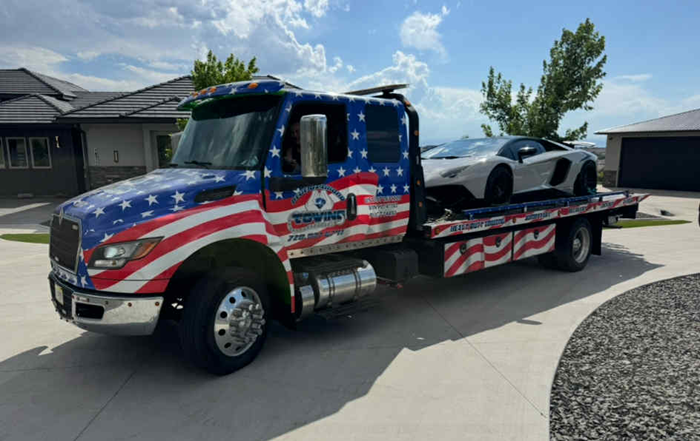 Tow truck with American flag design transporting a white Lamborghini on a sunny day.