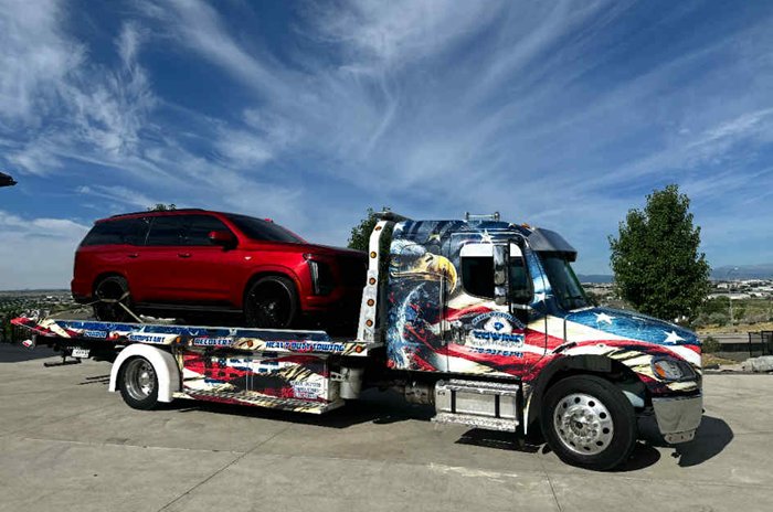 Tow truck with patriotic wrap carrying a red SUV on a sunny day.