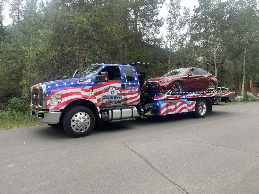 Tow truck with American flag design hauling a red car on a road near trees.