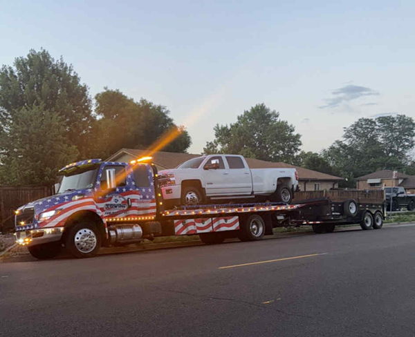 Tow truck with American flag paint job hauling a white pickup truck and a trailer on a street at dusk.