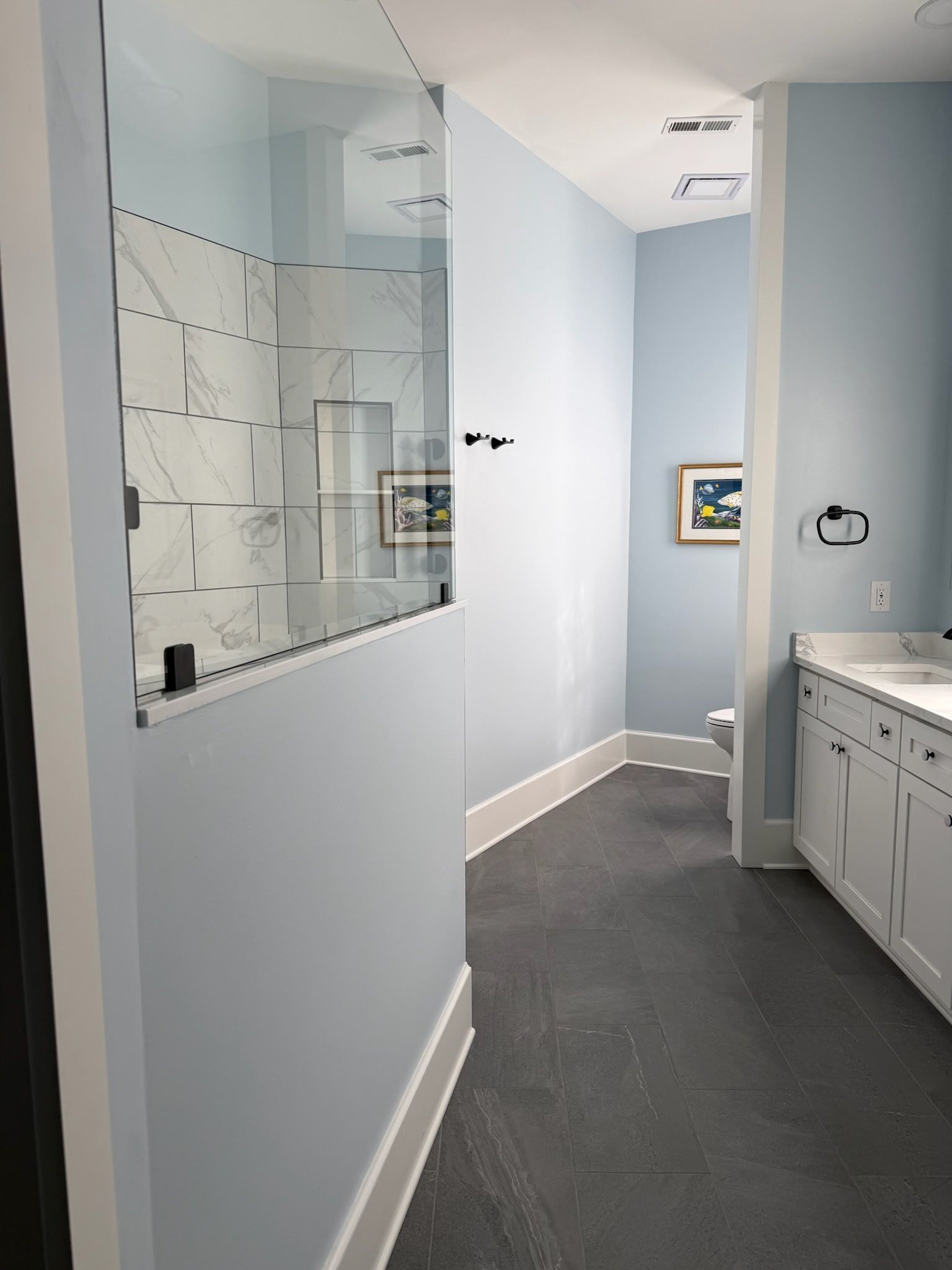 A bathroom with a half-wall glass shower, light blue walls, white cabinetry, and dark gray flooring.