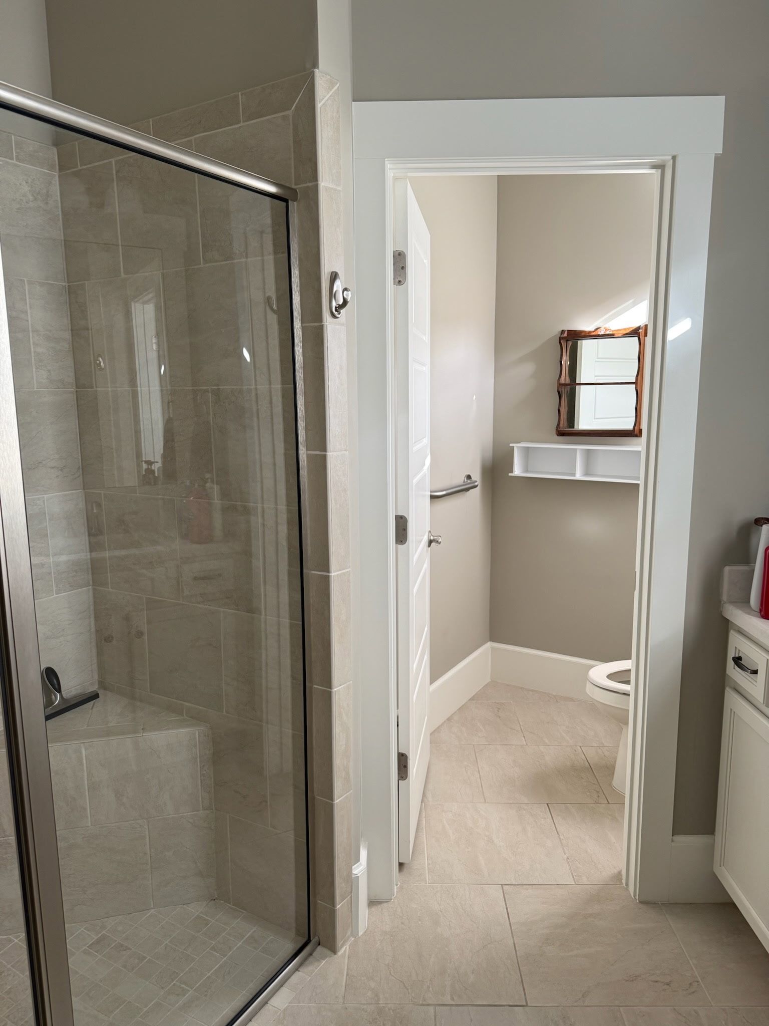 A glass shower with beige stone tiles next to a doorway leading to a small bathroom with a mirror and white shelves.