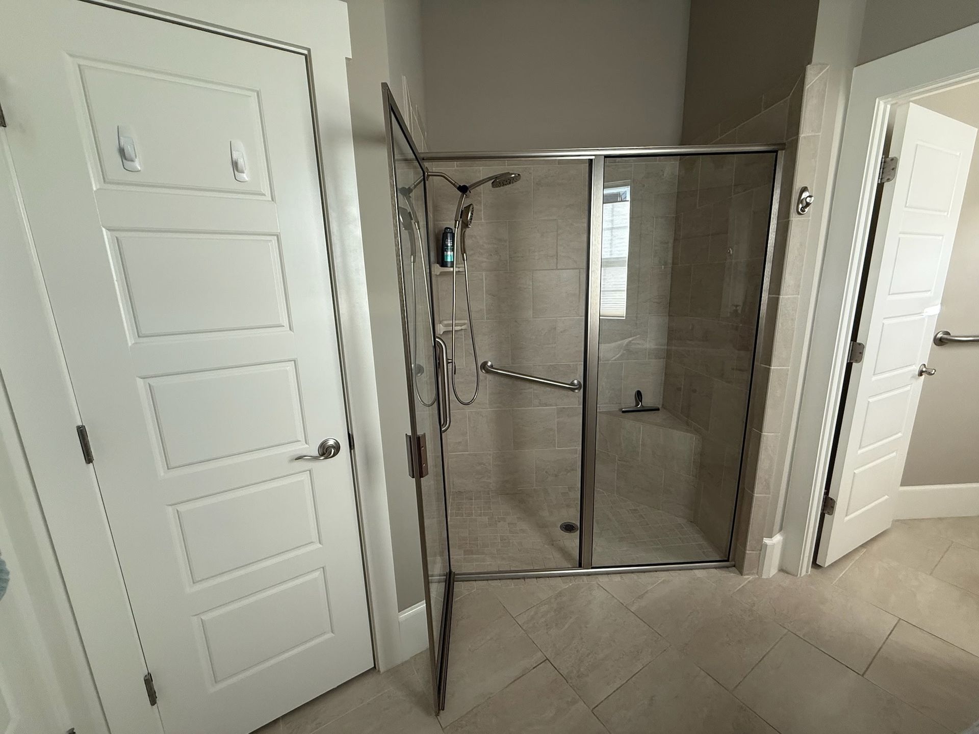 A bathroom interior featuring a walk-in glass shower with a bench, positioned between two white doors on tiled flooring.