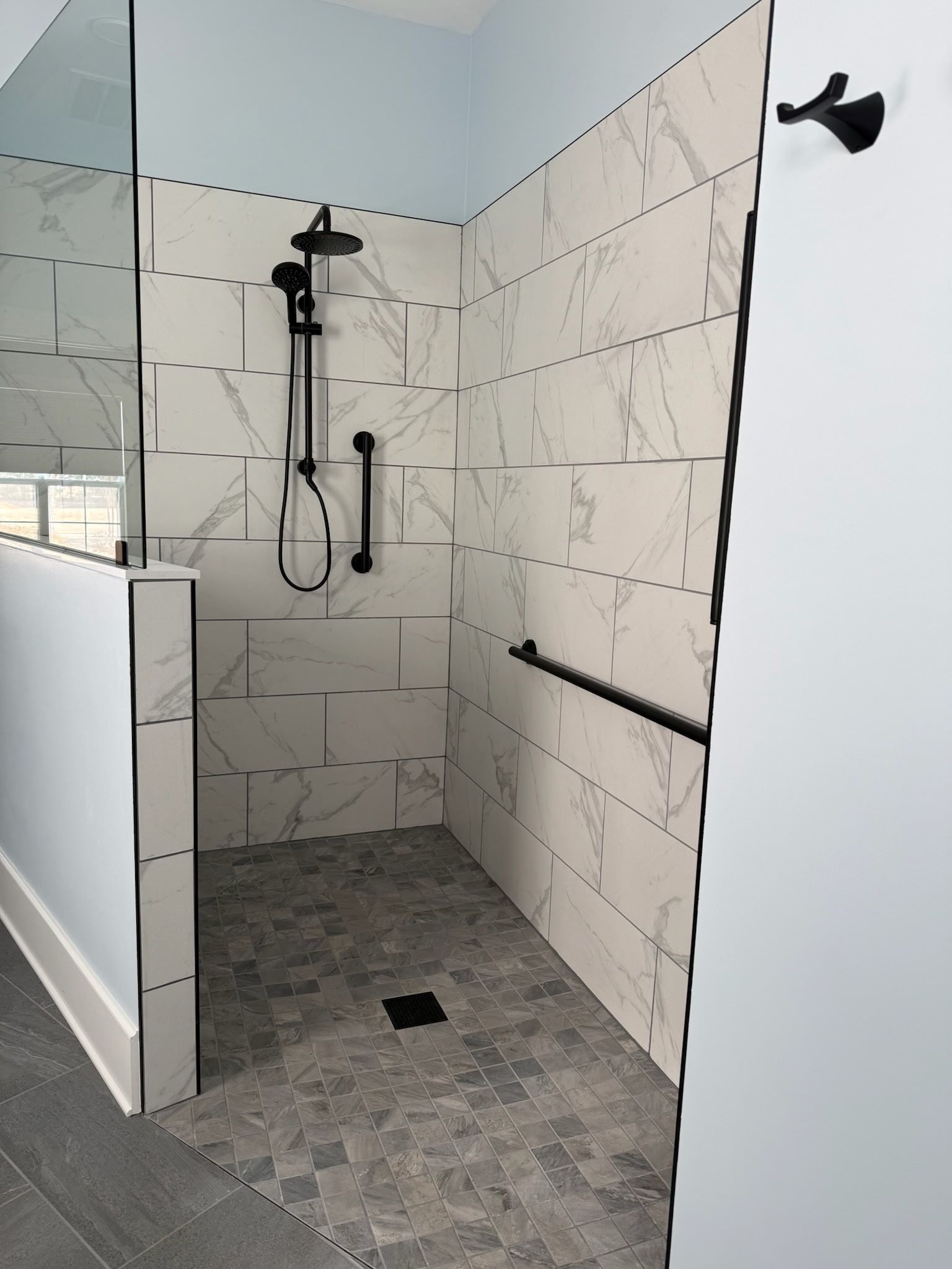 Walk-in shower with white marble-style tiled walls, black fixtures, grab bars, and grey stone-textured flooring.