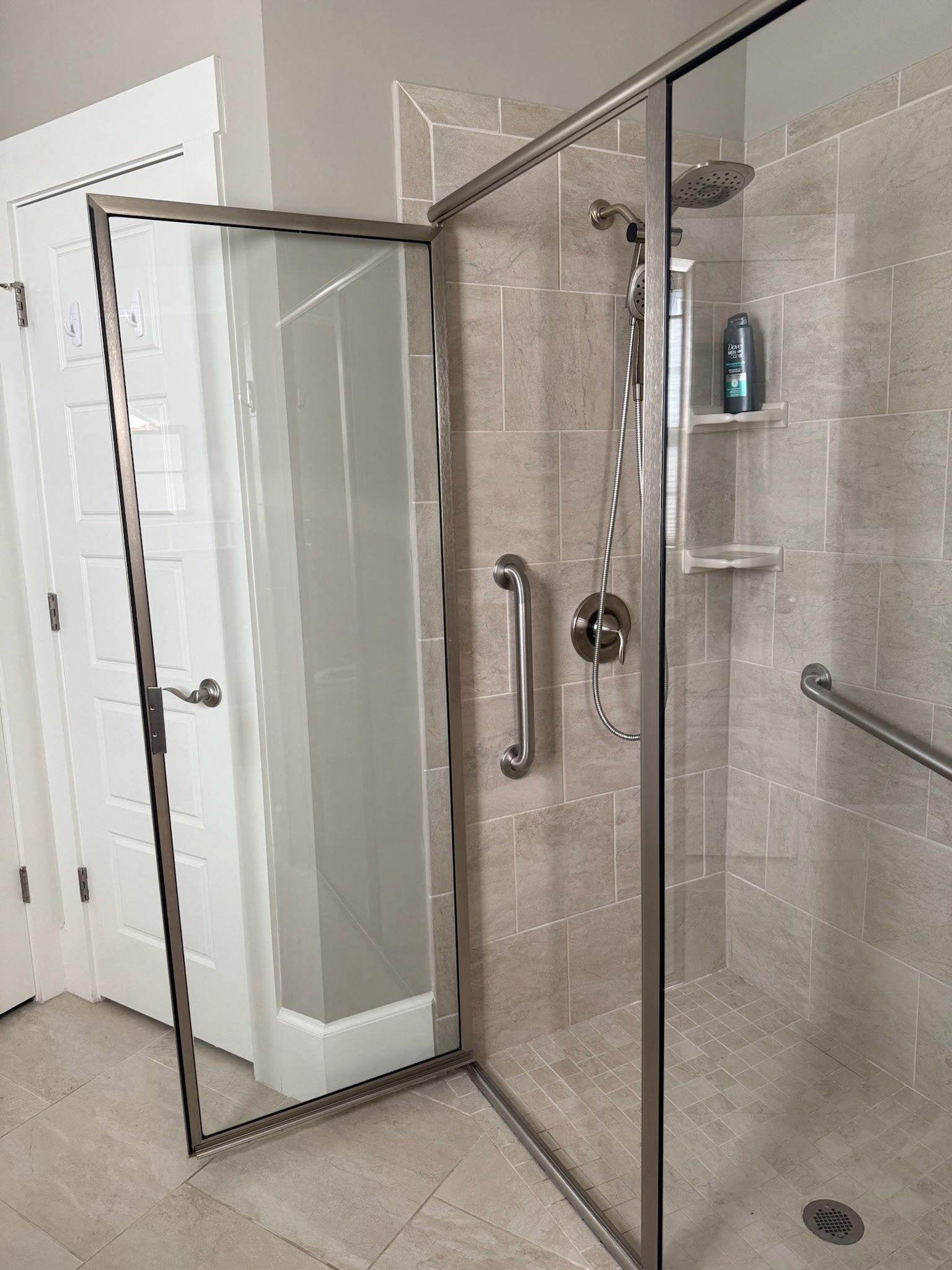 A glass-enclosed shower featuring tiled walls, two metal grab bars, a handheld shower head, and a built-in corner shelf.
