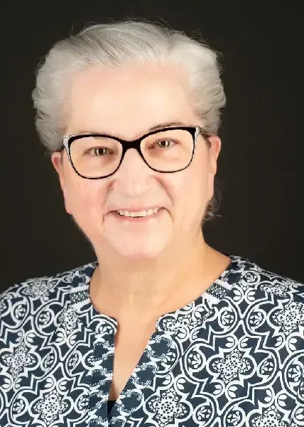 Portrait of a smiling person with short white hair, black glasses, and a patterned navy blouse on a dark background