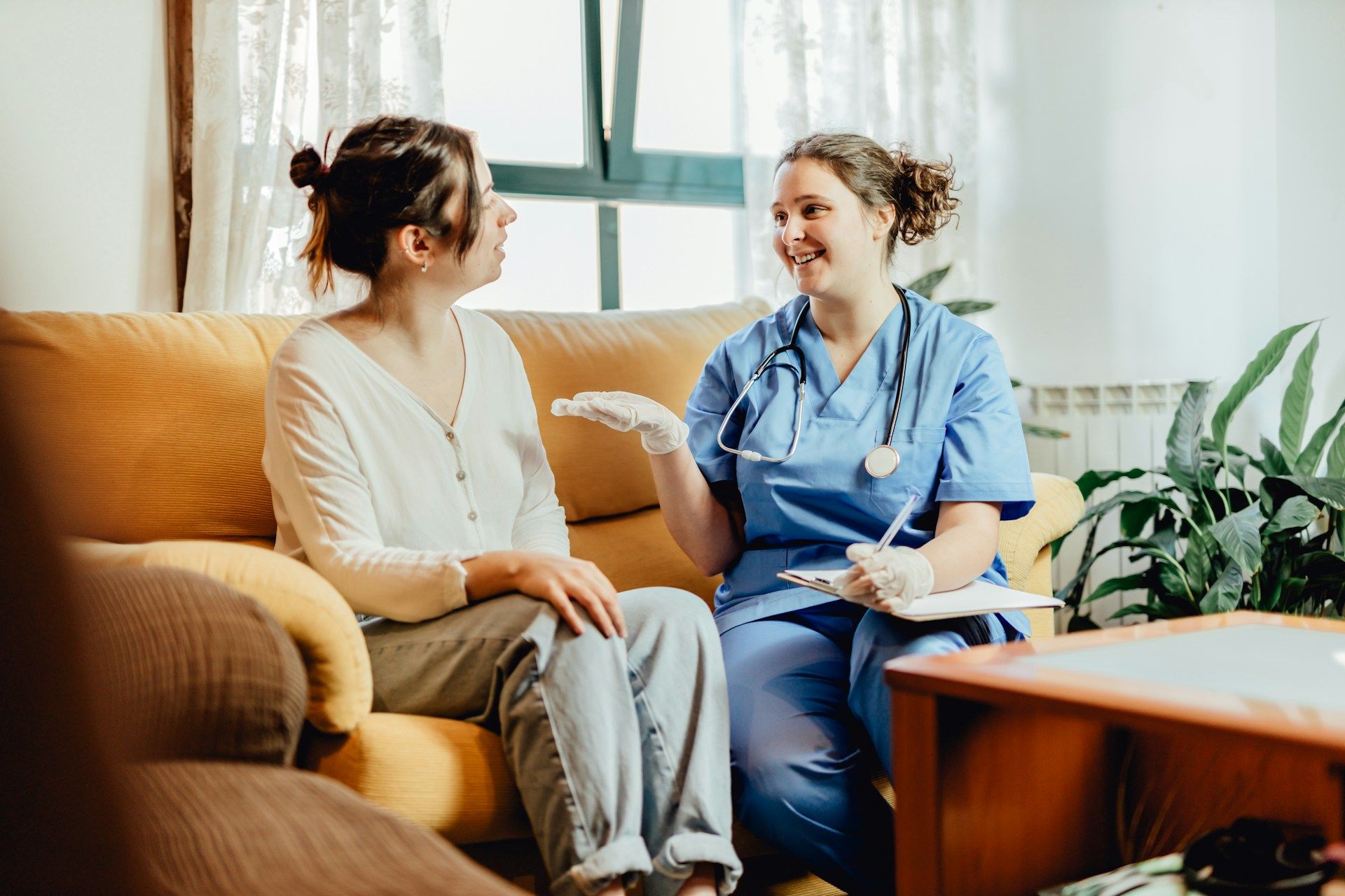 Two women talking on a couch in a bright living room, one in blue scrubs with a stethoscope