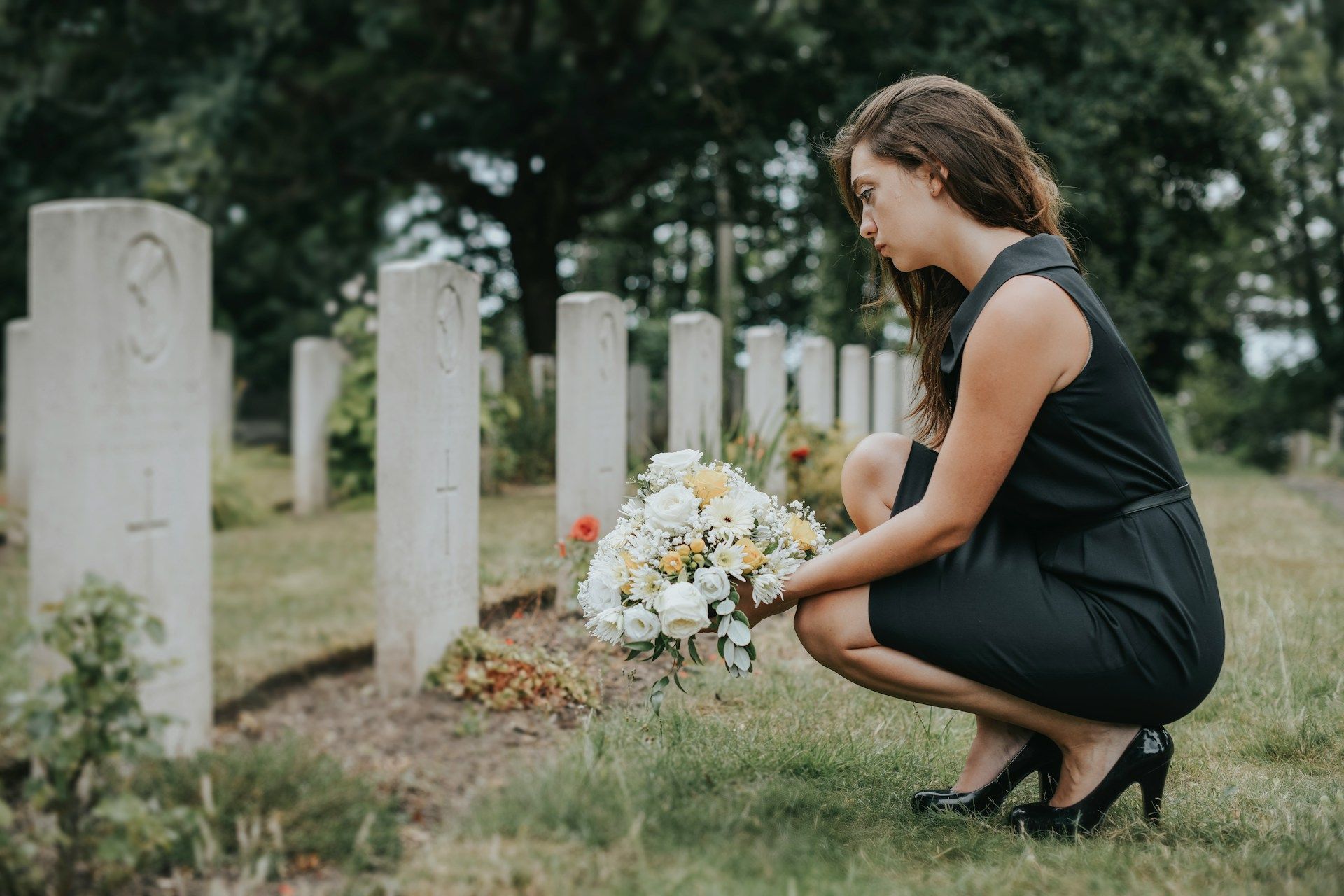Woman kneeling beside gravestones, placing flowers at a cemetery on a cloudy day