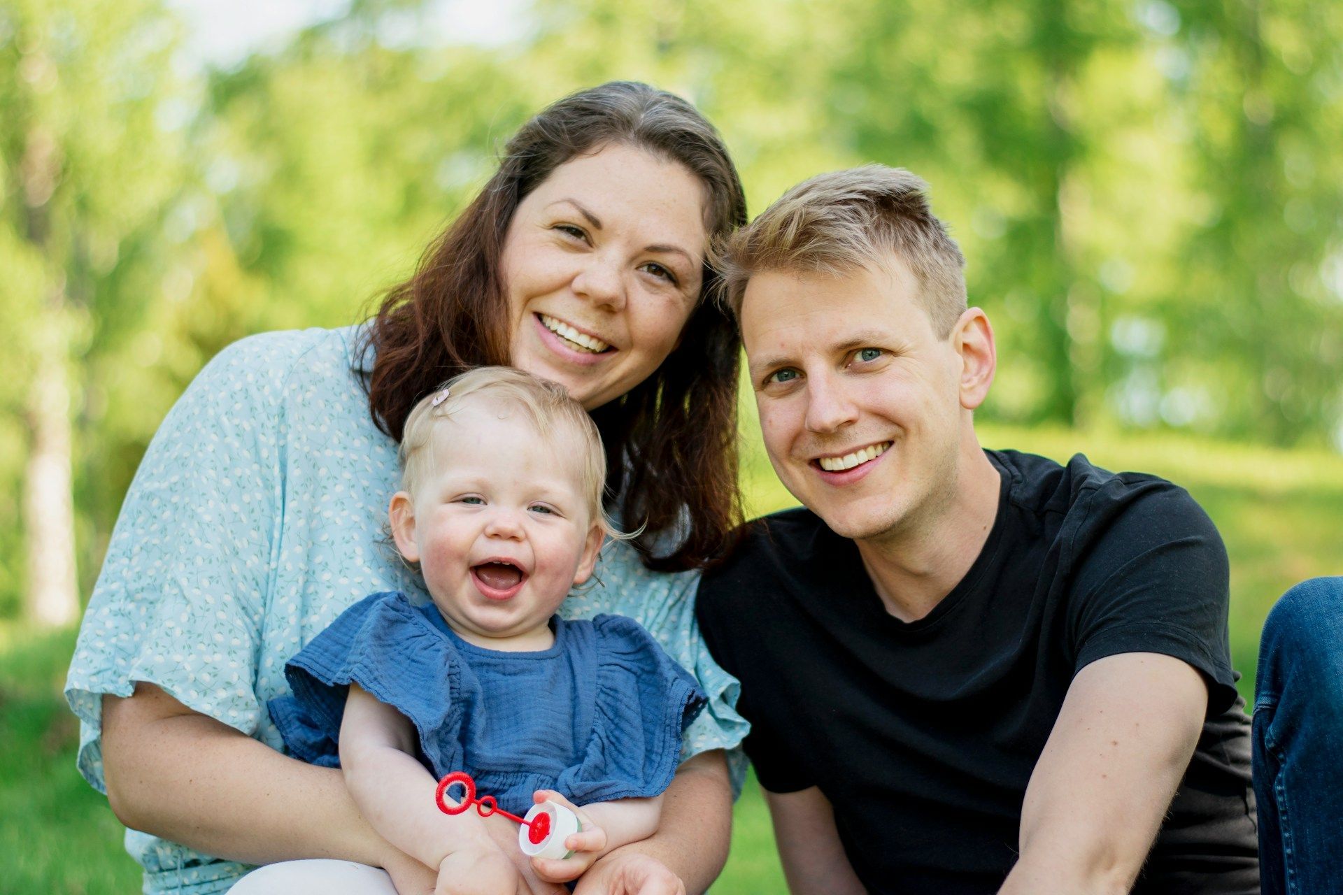 Smiling family seated outdoors in a green park, with an infant in the foreground.