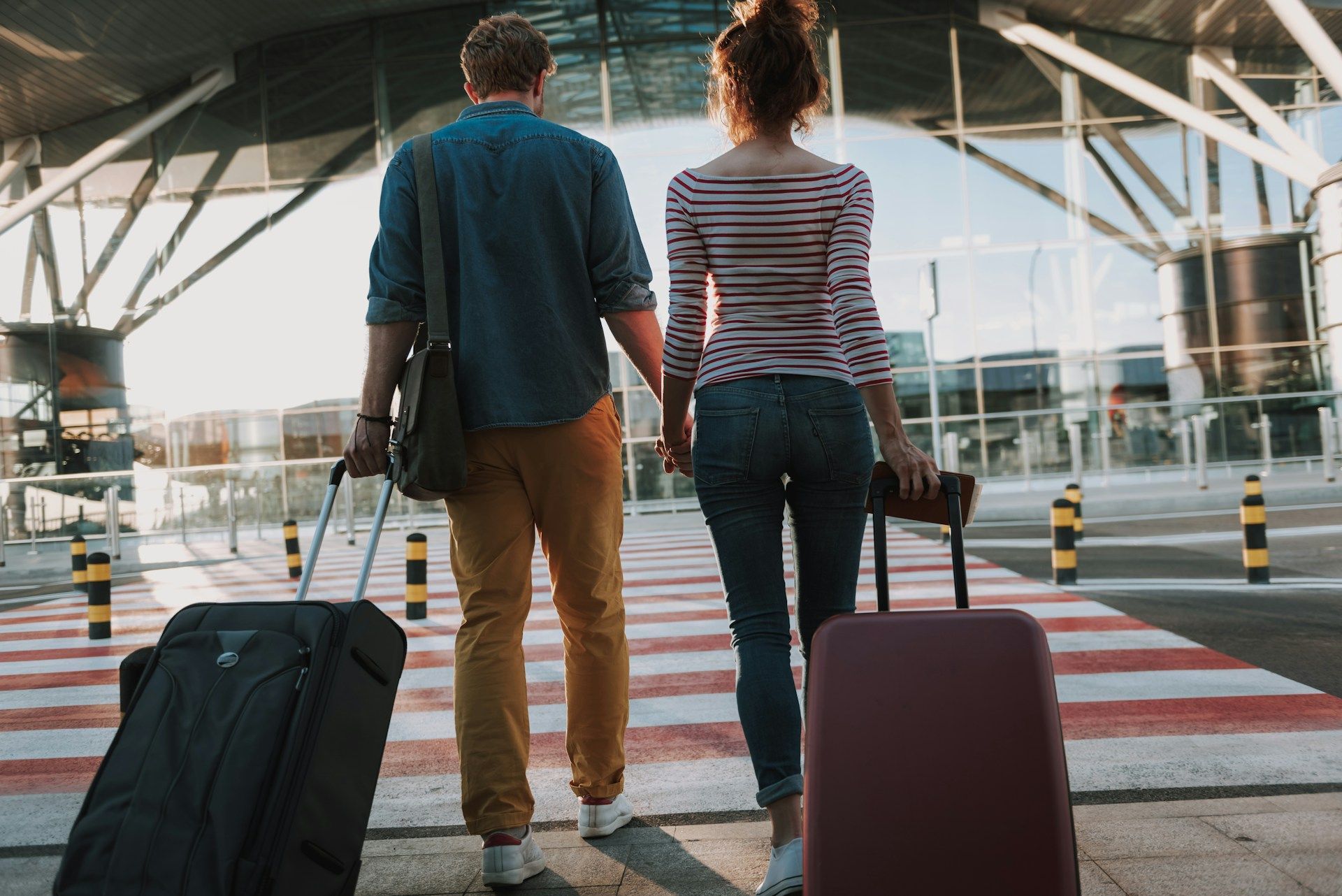 Two travelers with rolling suitcases walking across an airport crosswalk, seen from behind.