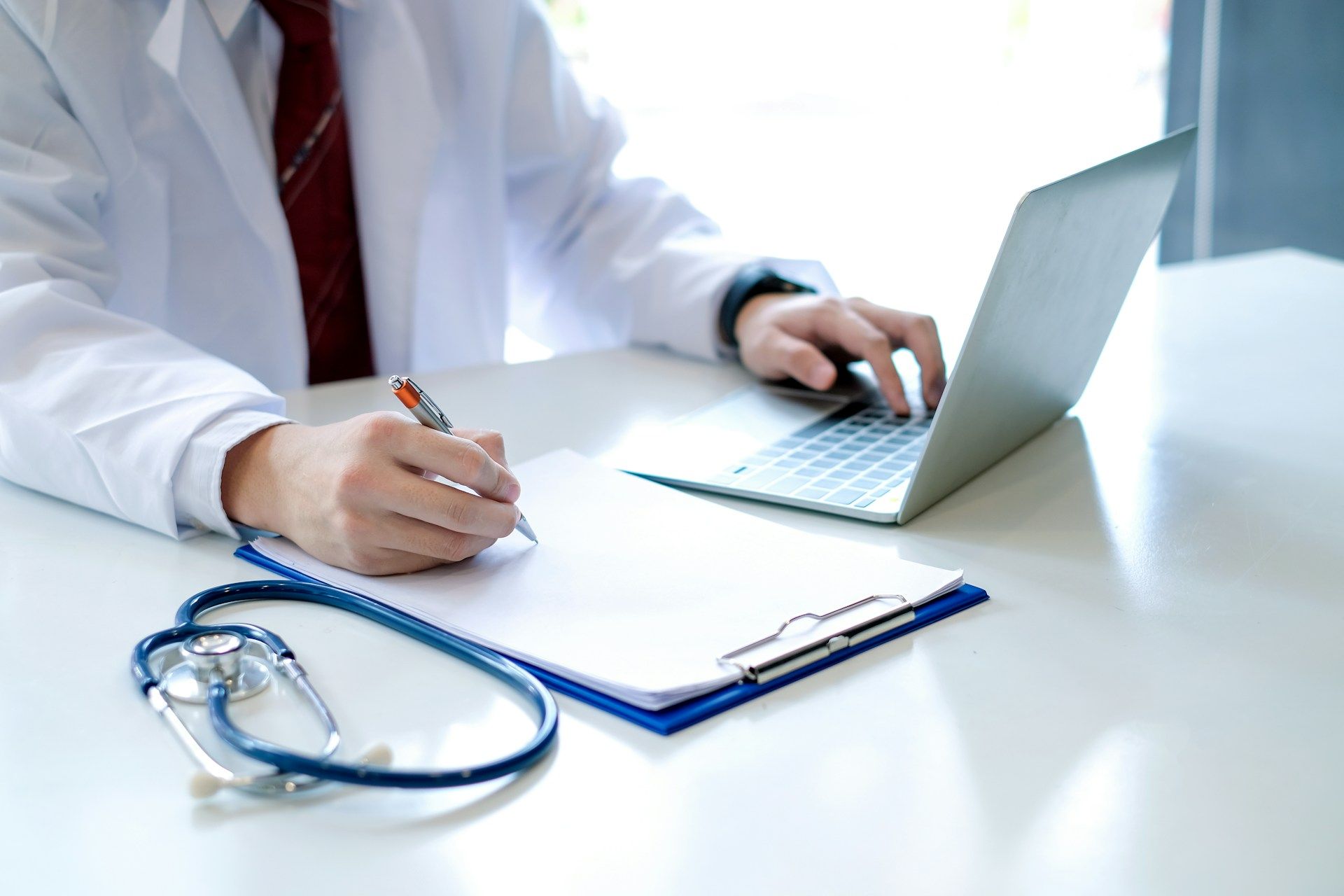 Doctor writing notes beside a laptop and stethoscope on a desk