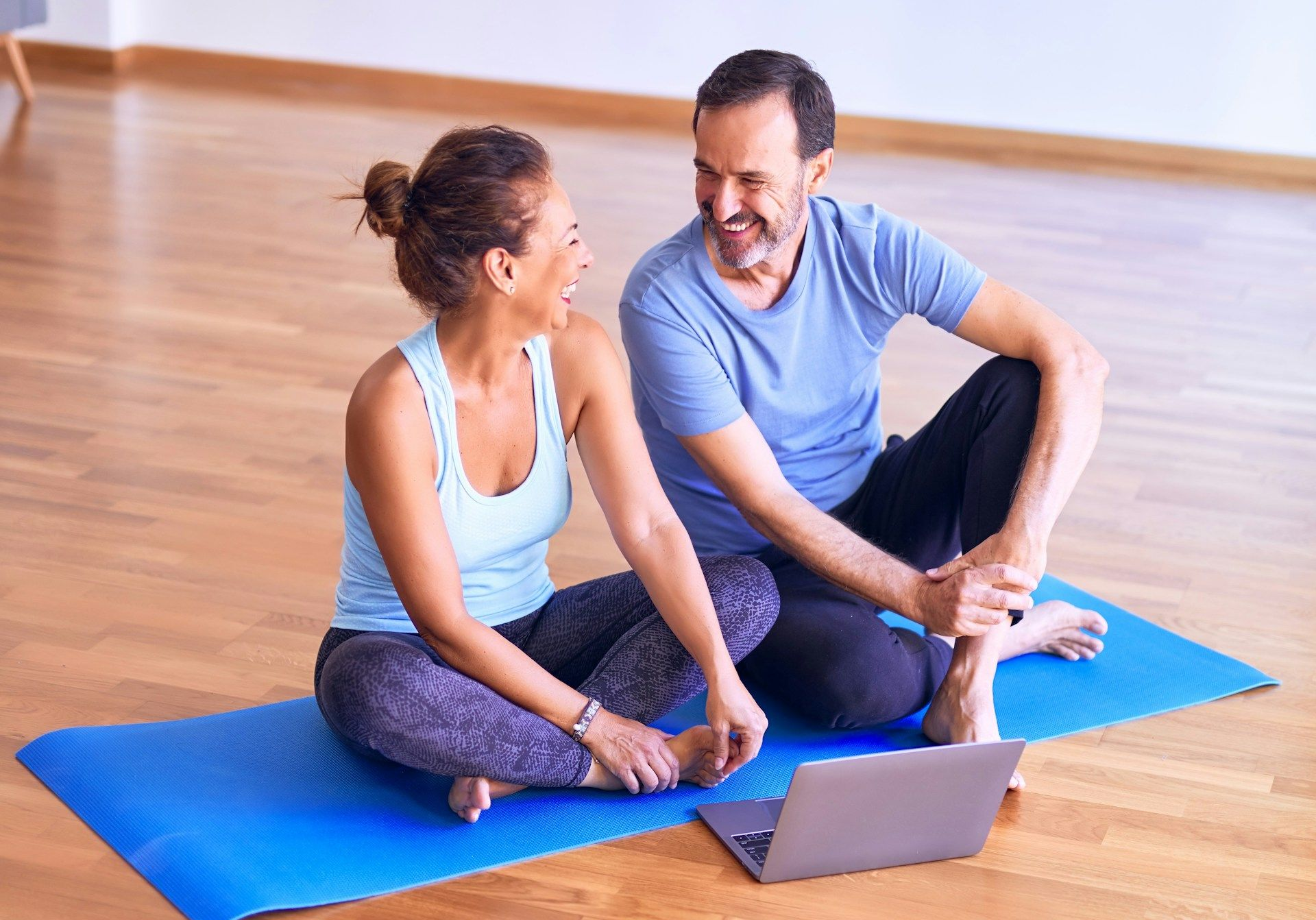 Two people stretching on blue yoga mats, smiling at a laptop in a bright studio