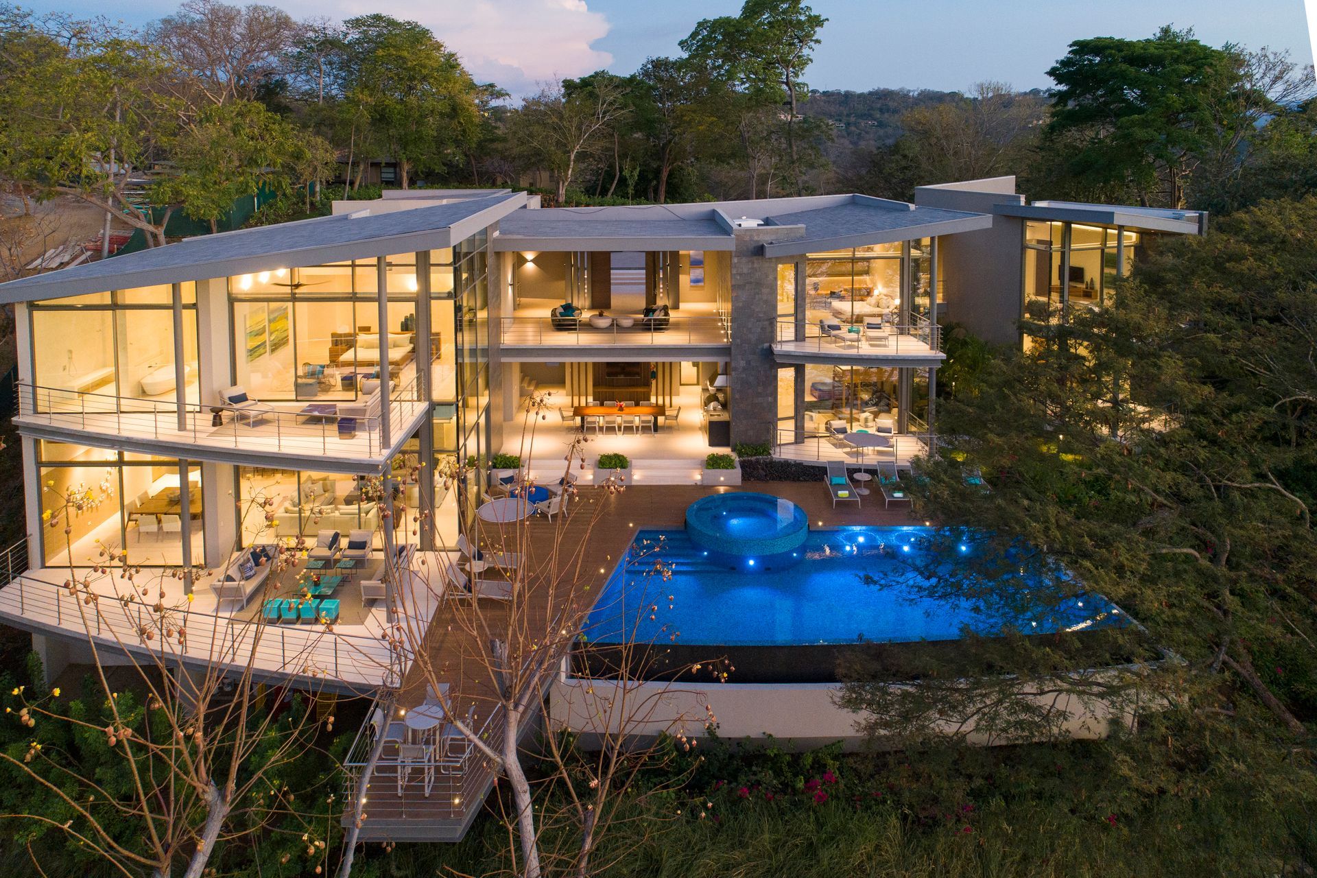 Modern multi-story house with a blue pool, glass walls, and surrounding greenery.