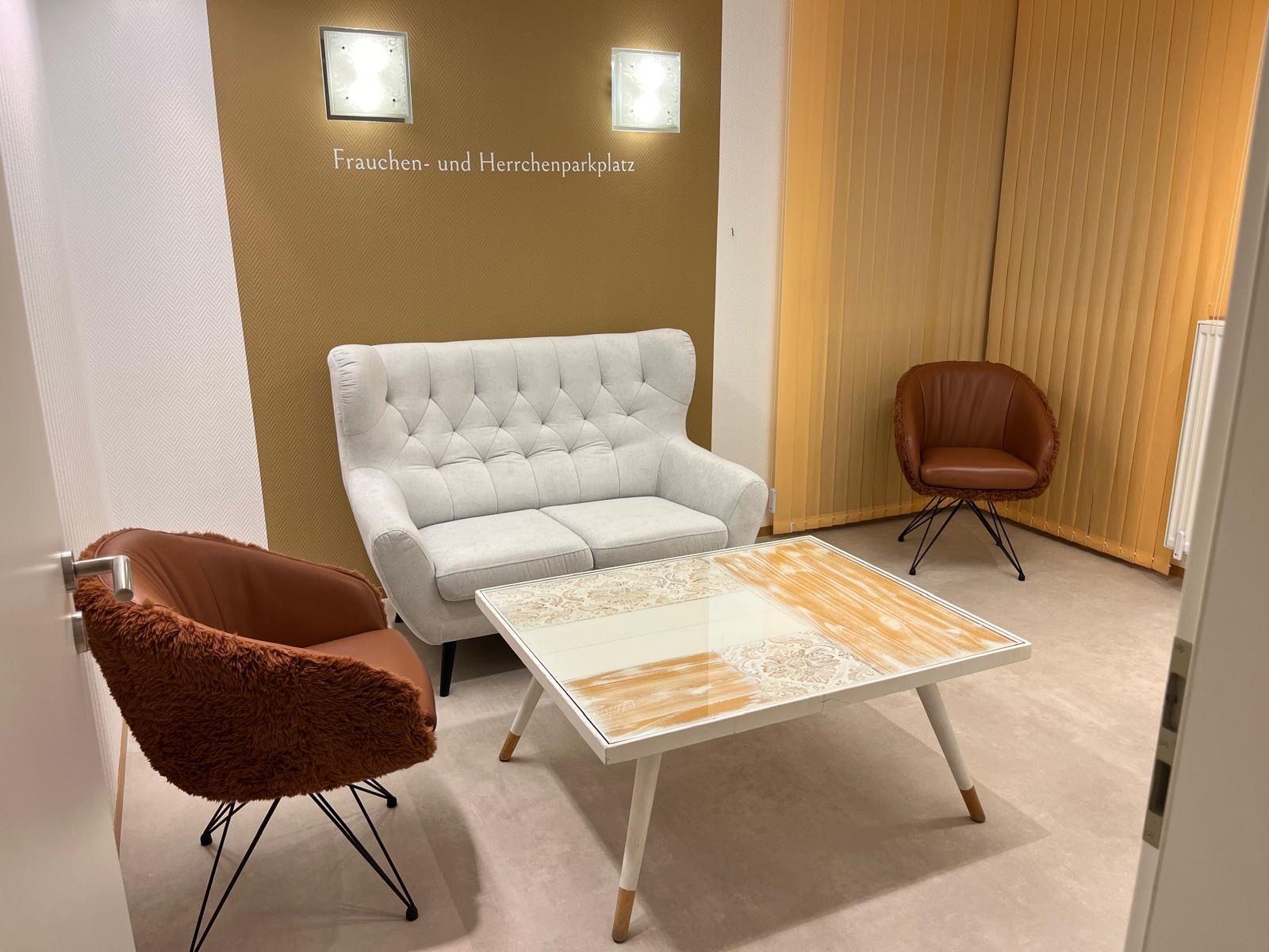 Gemütliches Wartezimmer mit weißem Sofa, zwei braunen Sesseln und einem Couchtisch. Goldene Akzentwand.