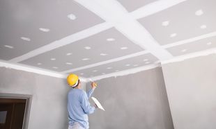 Worker in hard hat painting a white ceiling in a room under construction