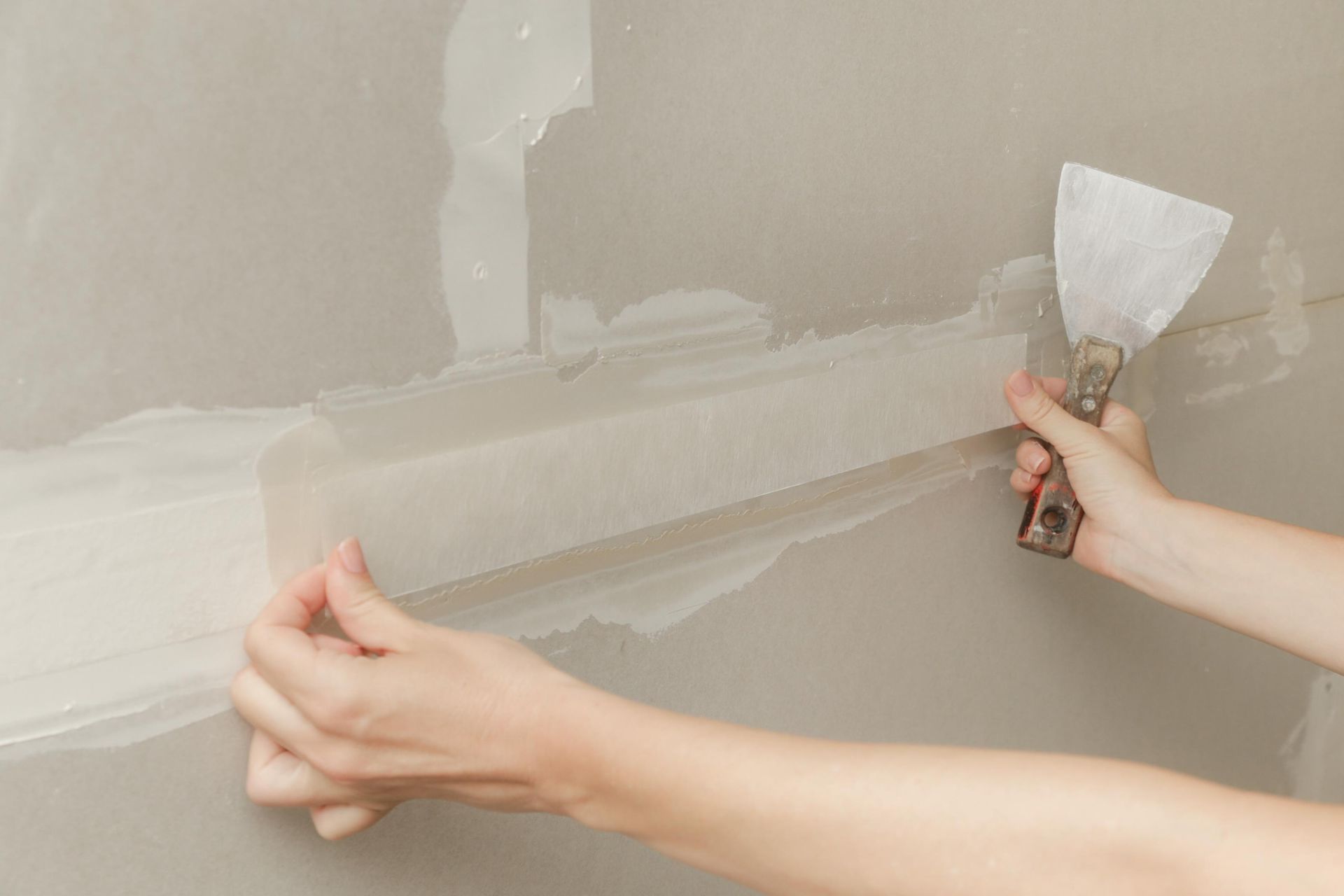 Hands smoothing drywall joint compound with a putty knife on a beige wall