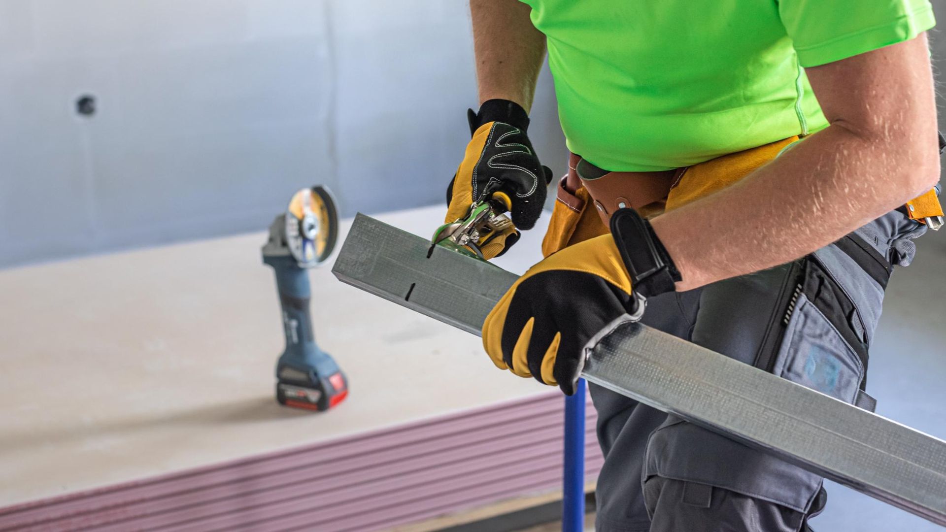 Worker measuring a metal beam with a tape measure, wearing gloves and a bright green shirt.