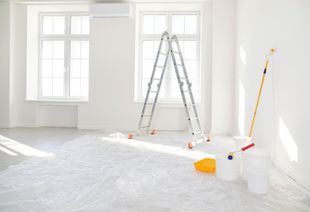 Bright empty room with ladders, paint tools, and covered floor during renovation