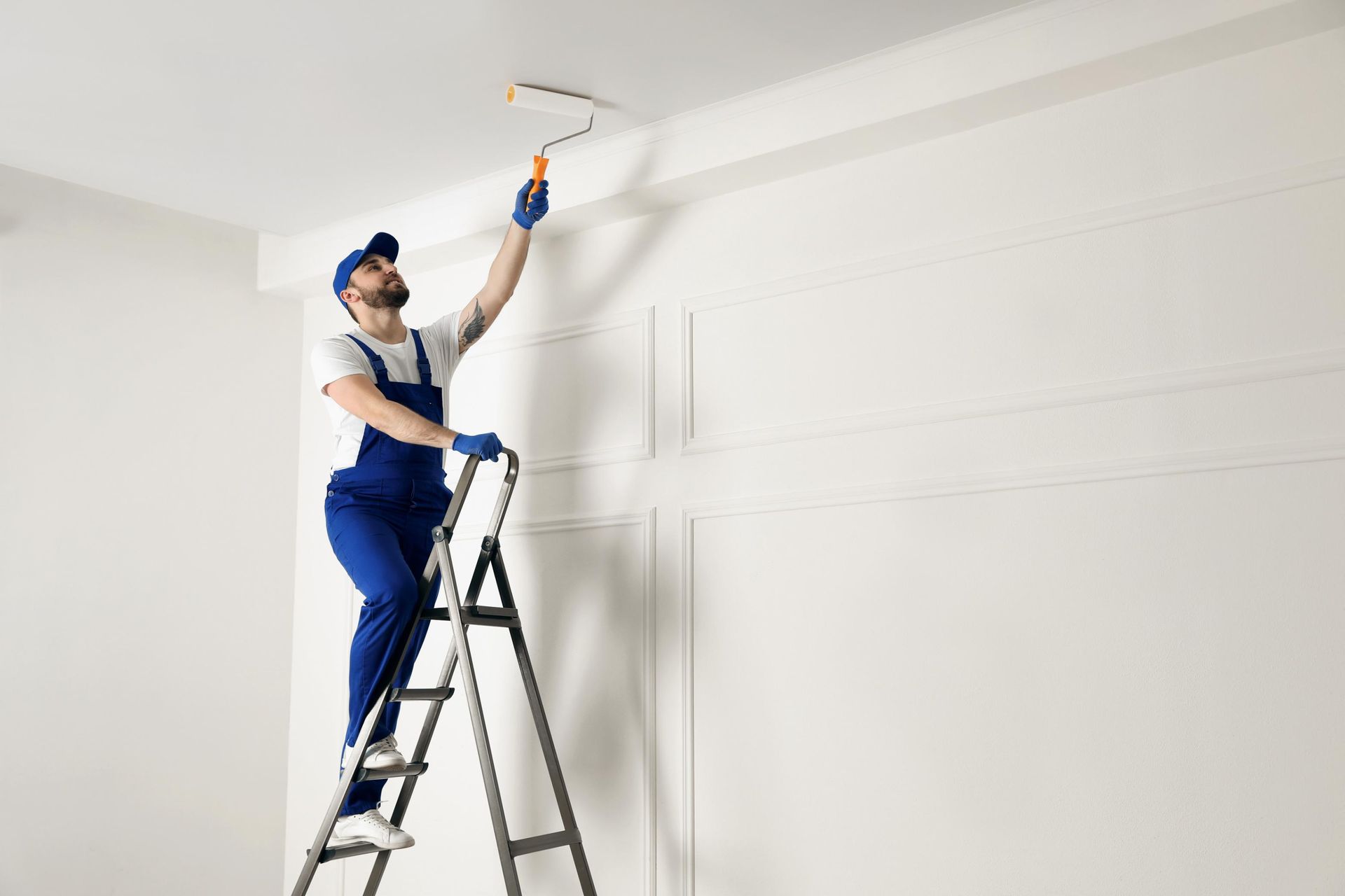Person on a ladder painting a white ceiling with a paint roller in a bright room