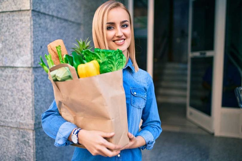 Smiling woman in blue shirt holding a grocery bag with fresh produce outside a building