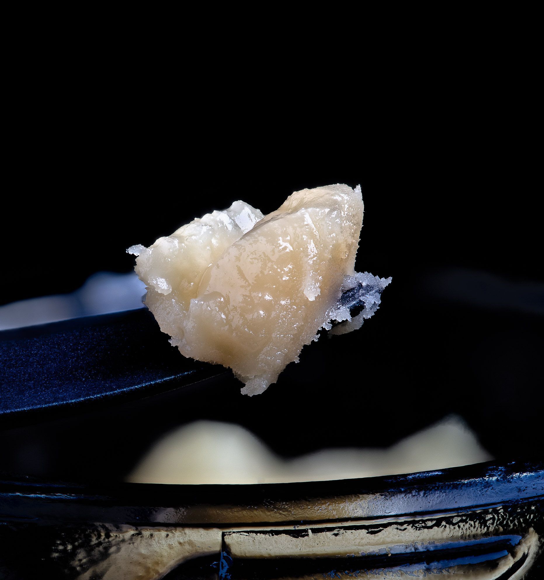 Dab of cannabis concentrate on a black tool, held over a black container. The concentrate is light brown, crystalline.
