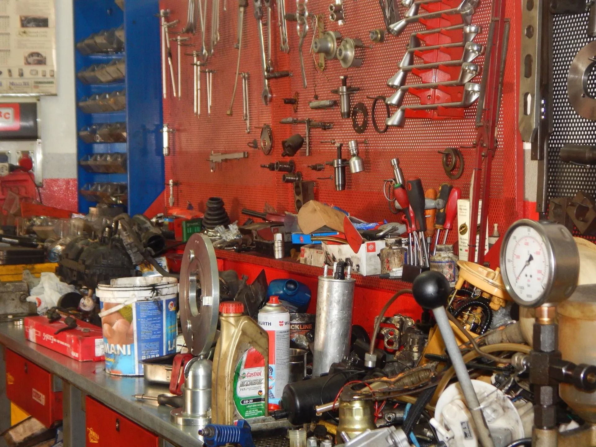 Cluttered mechanic’s workbench with tools, spray cans, and gauges on a red pegboard wall