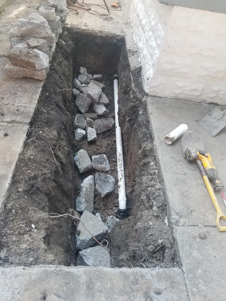 A trench dug in concrete, containing a PVC pipe and rocks. A sledgehammer and pipe are nearby.