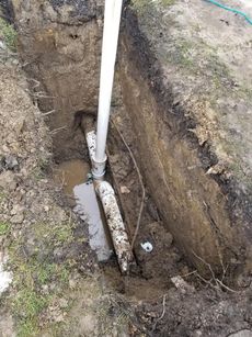 Trench in soil with white PVC pipe and connections, next to a vertical pipe.