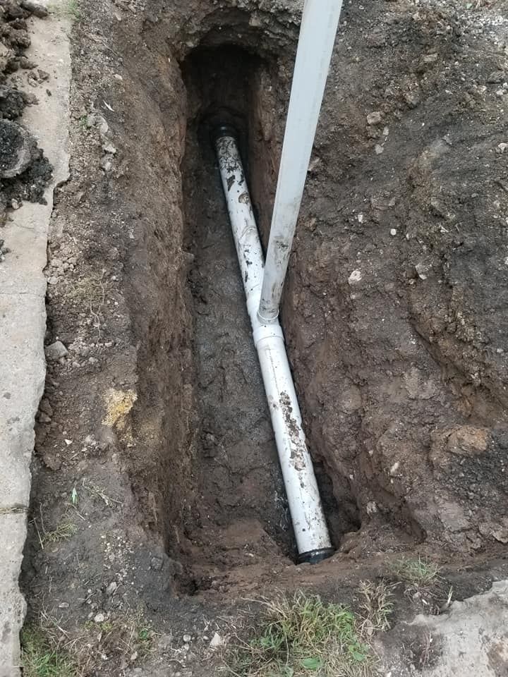 Trench with white PVC pipes; Y-shaped pipe, dirt, next to a concrete curb.