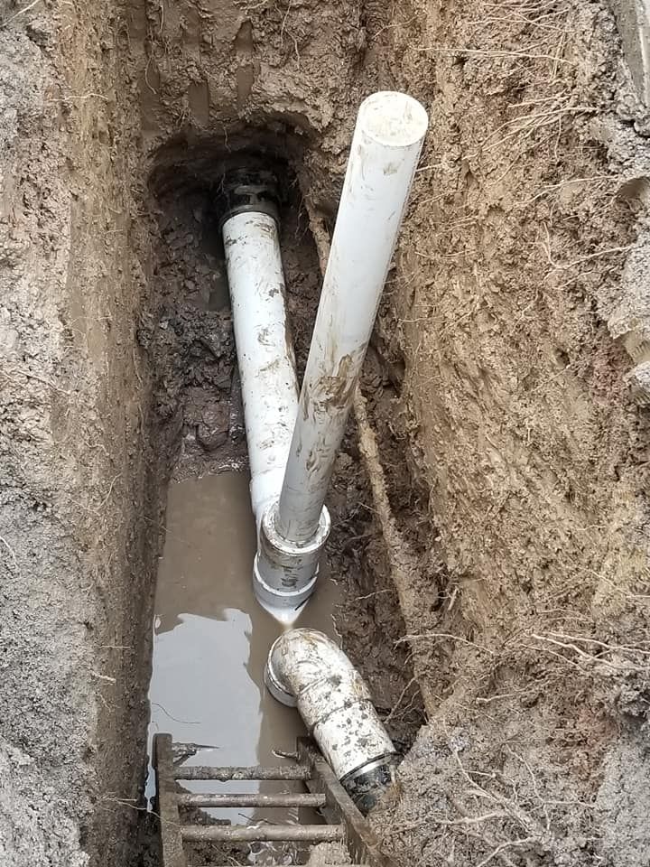 Pipes in a trench, possibly a repair. White and gray pipes visible, surrounding wet soil.