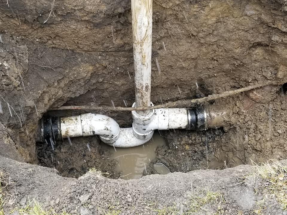 Plumbing repair in an excavated pit: white pipes, a curved trap, water, and brown soil.