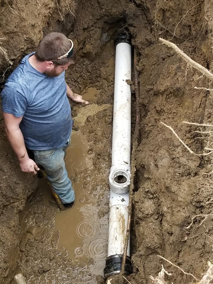 Man in a trench examining a vertical white pipe with a T-fitting, surrounded by wet dirt.