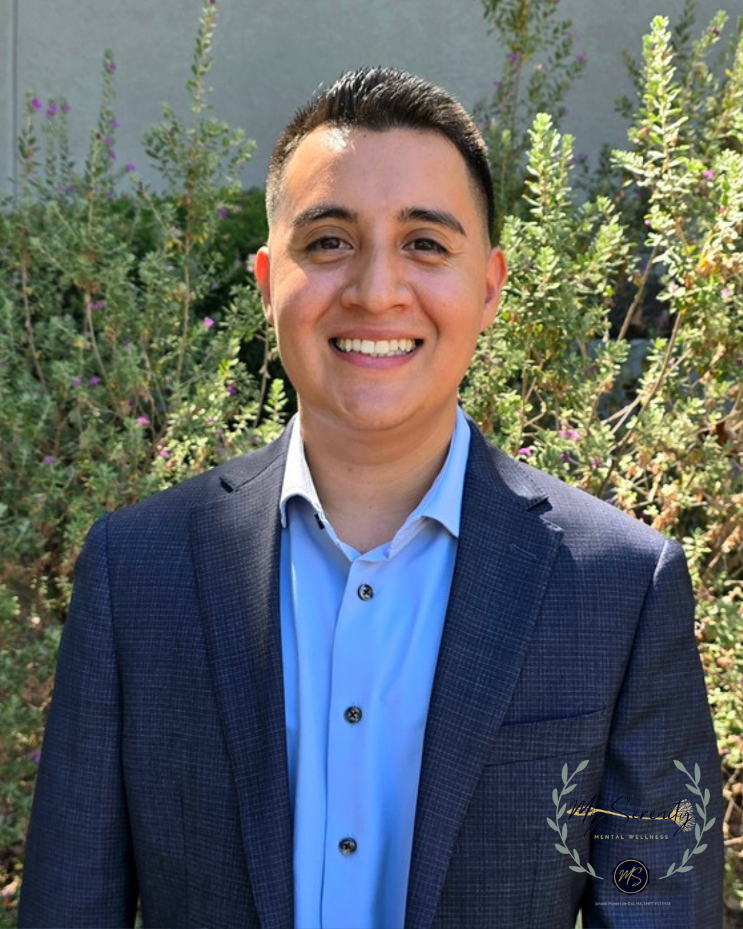 Smiling person in a navy blazer and blue shirt standing outdoors in front of greenery.