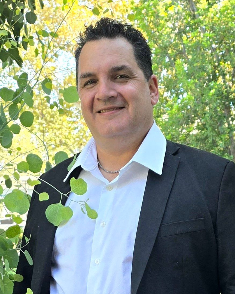 Smiling person in a black top standing outdoors in front of green leaves.