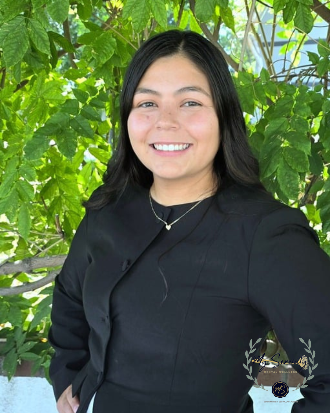 Smiling person in a black top standing outdoors in front of green leaves.