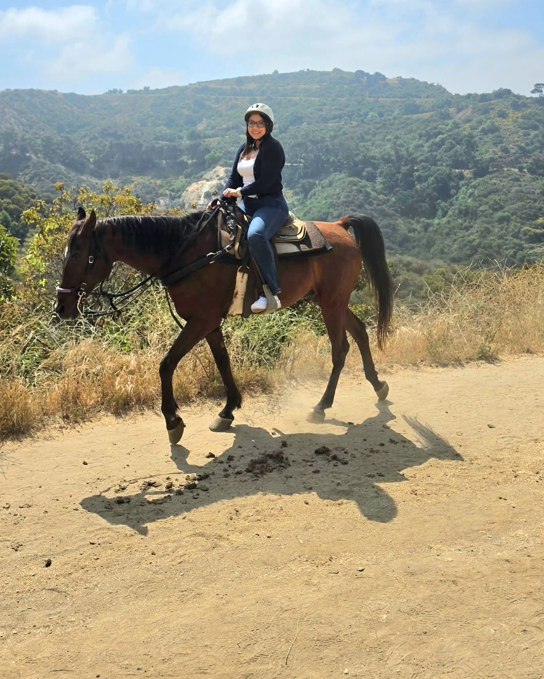 Person riding a brown horse on a dusty trail with green hills in the background