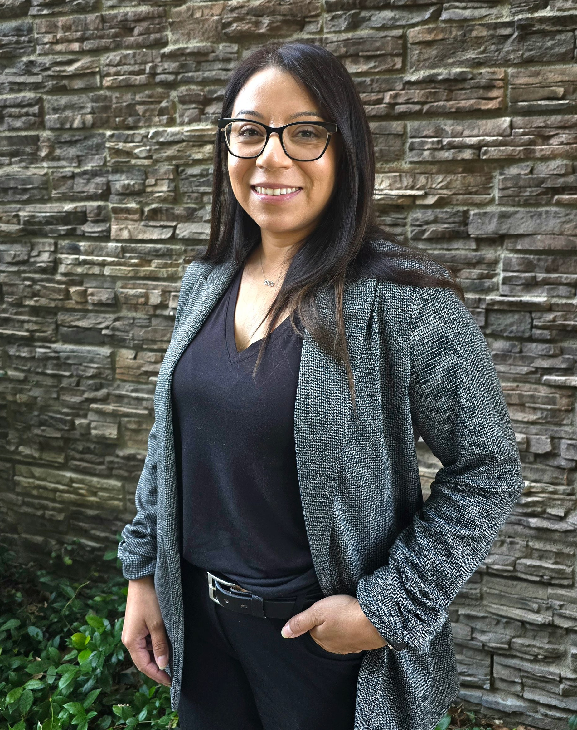 Smiling person in a black top standing outdoors in front of green leaves.