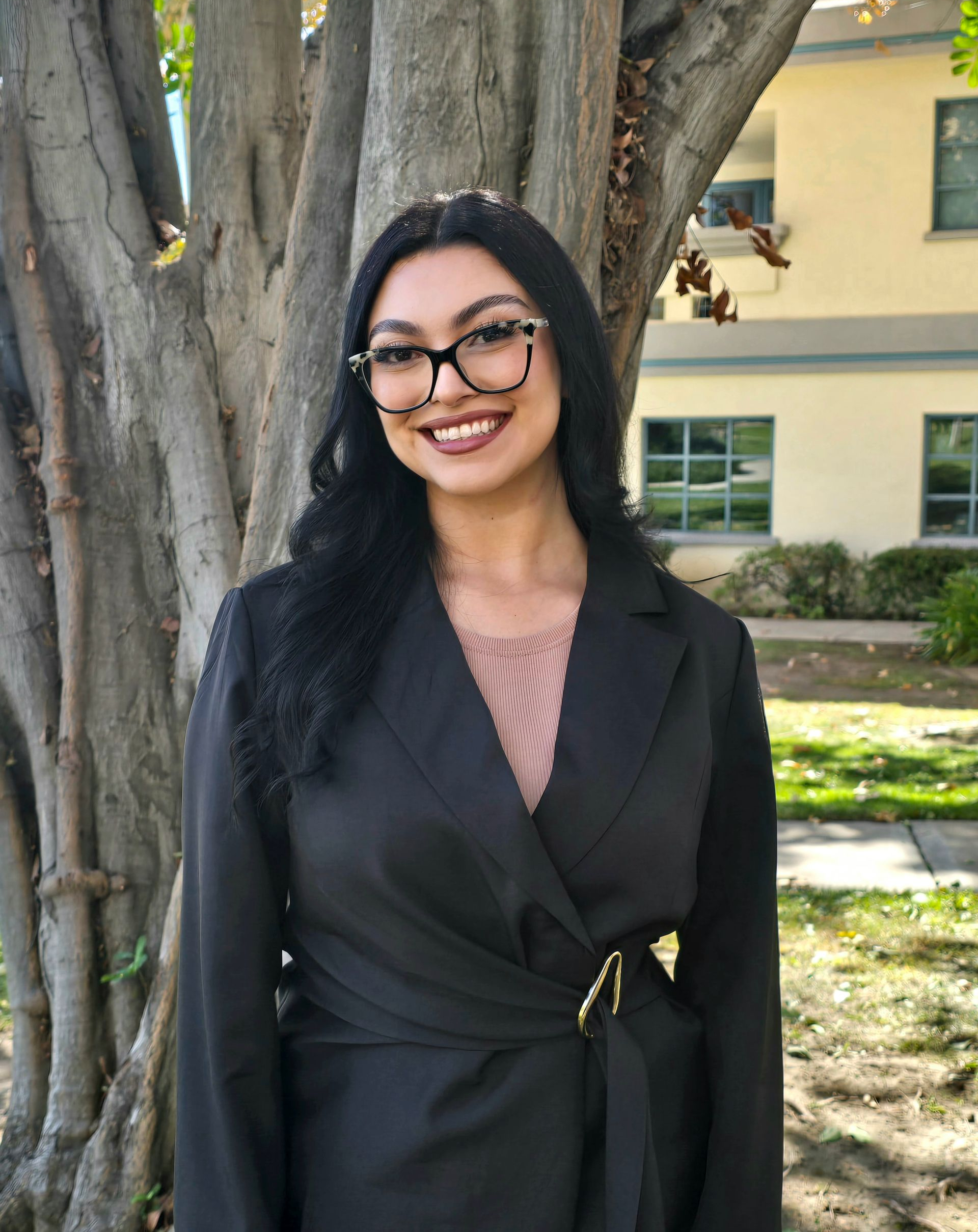 Smiling person in a black top standing outdoors in front of green leaves.