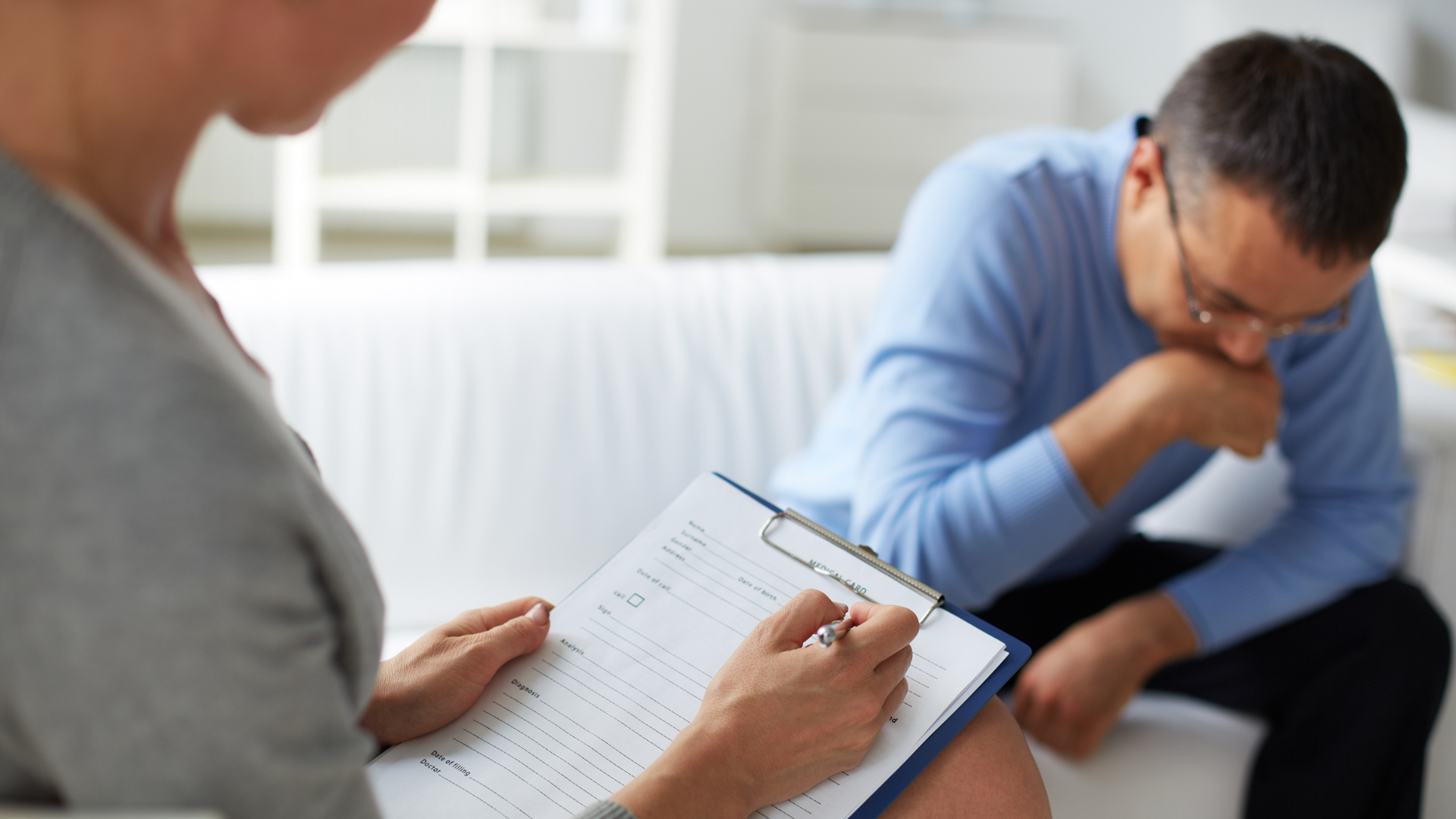 A man is sitting on a couch while a woman writes on a clipboard.