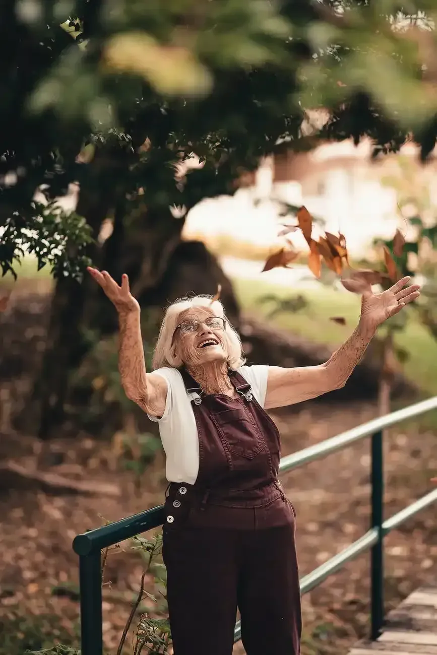 An elderly woman is throwing leaves in the air while standing next to a railing.