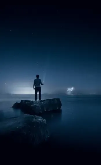 A man is standing on a rock overlooking the ocean at night.