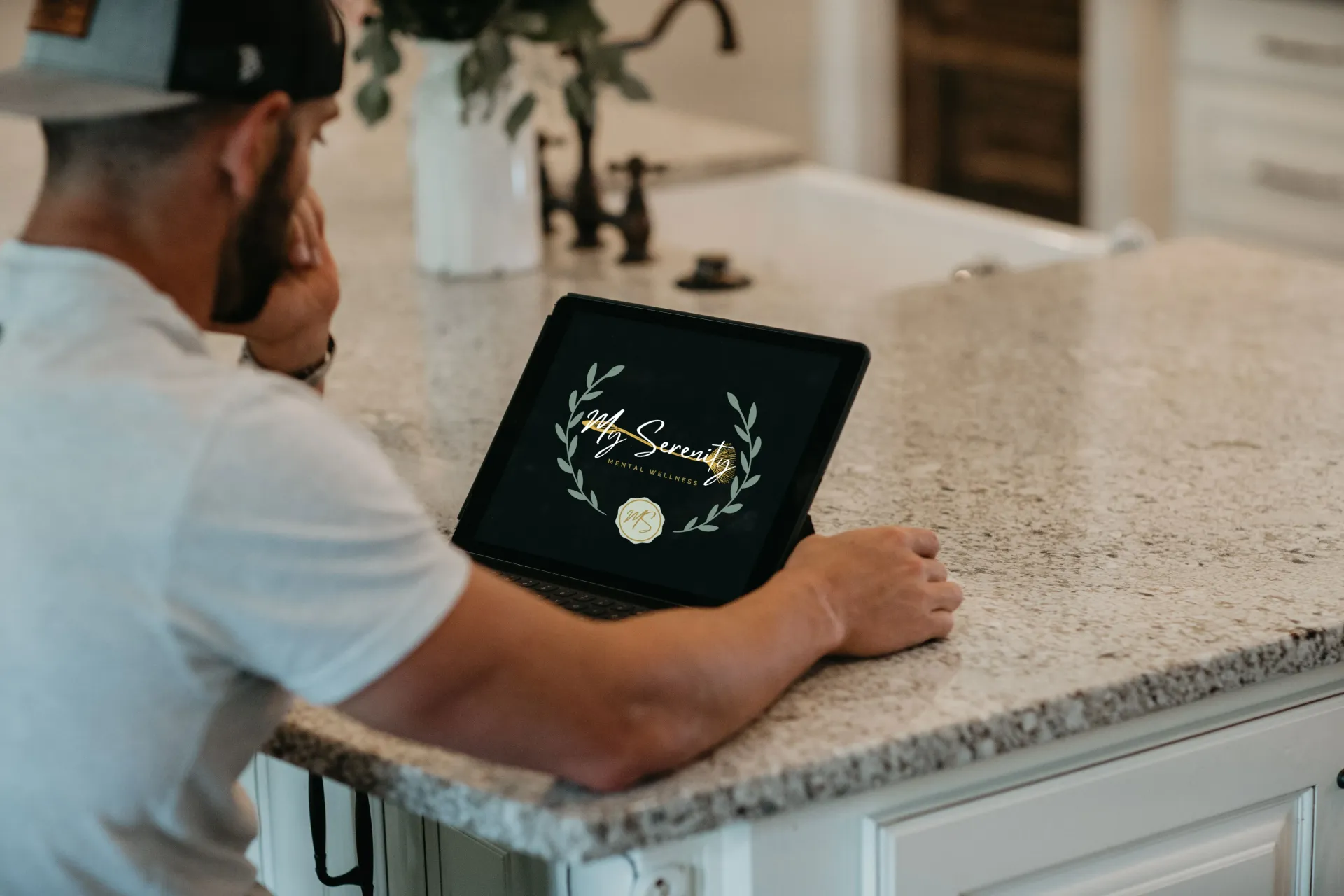 A man is sitting at a kitchen counter using a tablet computer.