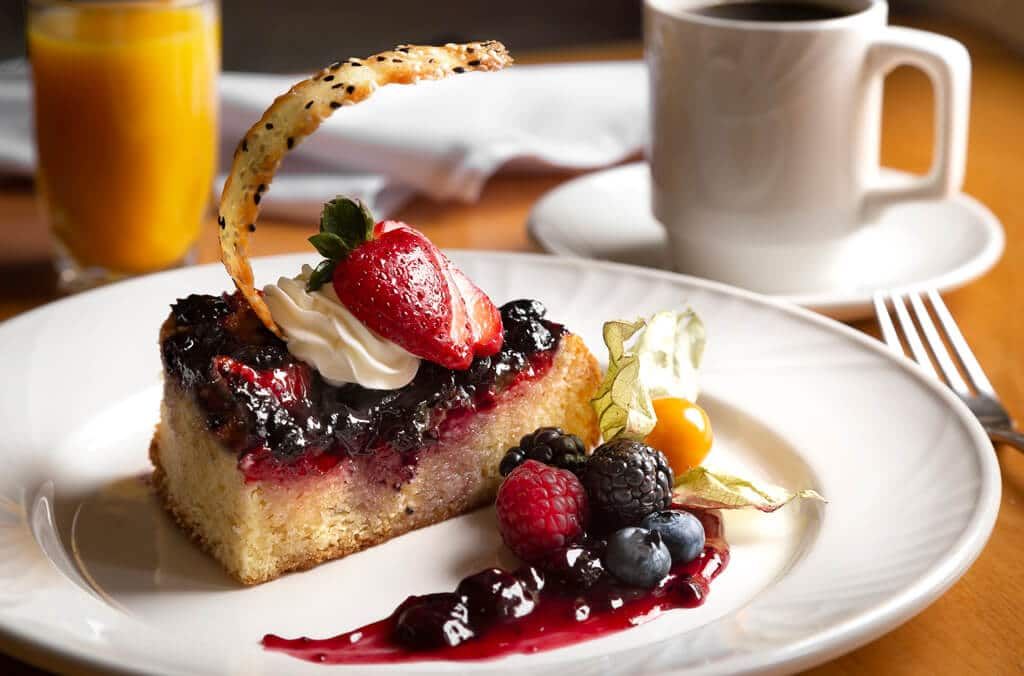 A white plate topped with a piece of cake and berries next to a cup of coffee