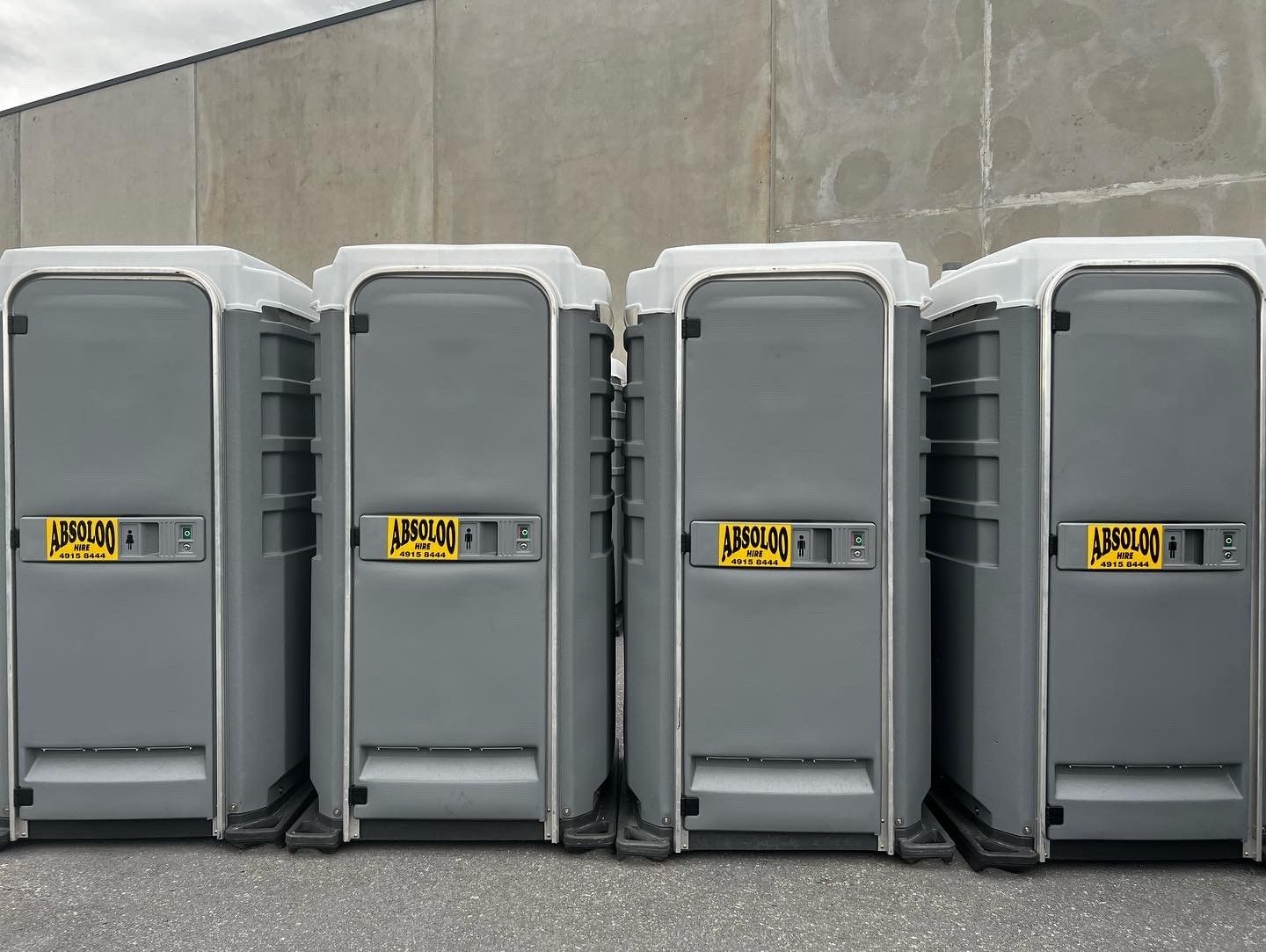 Four gray portable toilets lined up against a light gray wall — Absoloo Hire Pty Ltd In Morisset, NSW