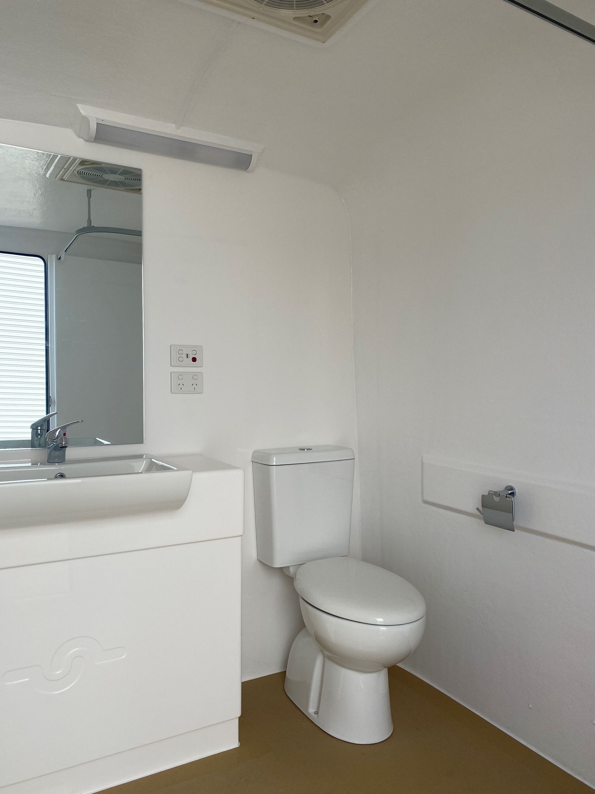 A small, white bathroom with a toilet, sink, and mirror. Brown flooring and white walls — Absoloo Hire Pty Ltd In Morisset, NSW